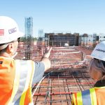 Twee bouwprofessionals met helm bespreken voortgang van een nieuwbouwproject op een bouwplaats met wapening en leidingen