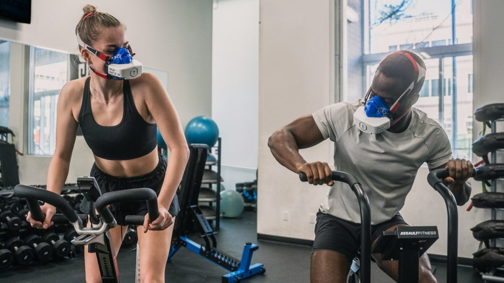 Sporters op airbike in de gym met trainingsmasker tijdens intensieve zuurstoftraining voor verbetering van uithoudingsvermogen